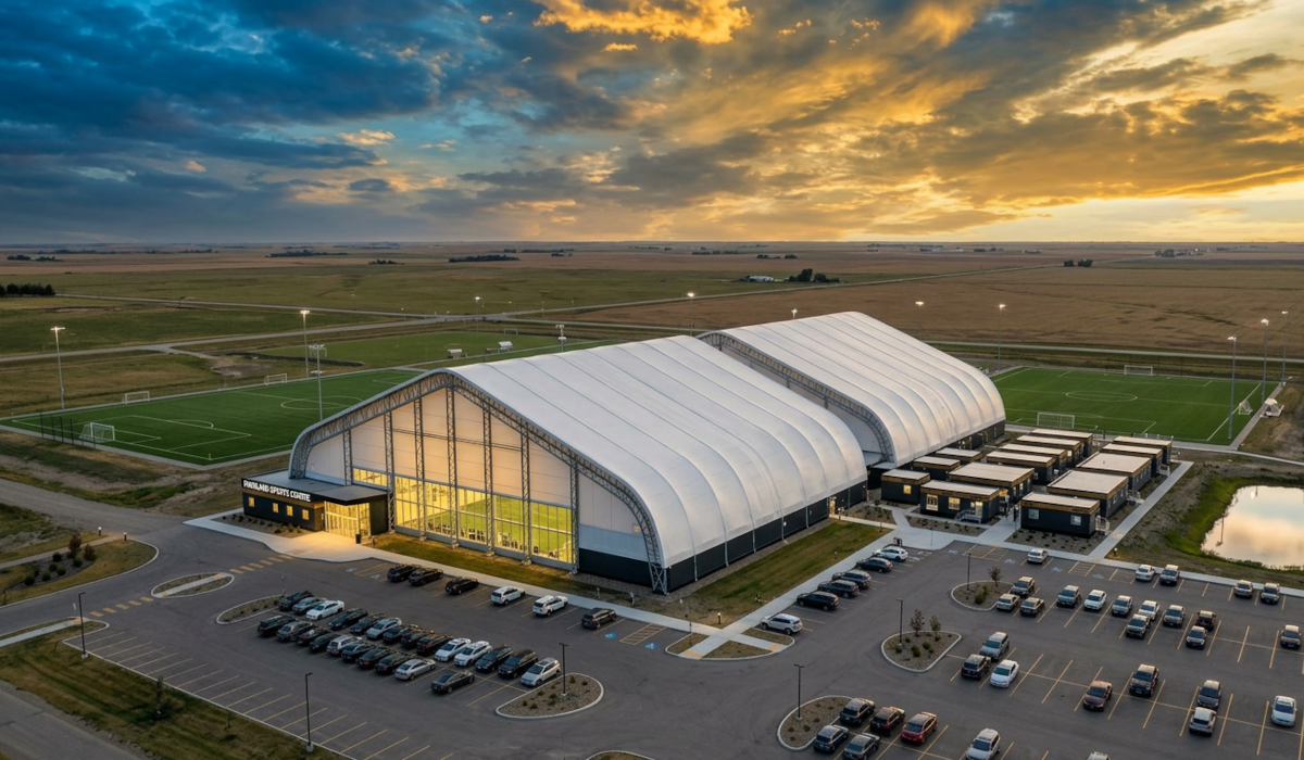 Aerial architectural rendering of indoor sports facility complex on flat Alberta prairie, two large fabric-covered double truss shelter structures side by side as main buildings with translucent panels glowing warmly, smaller modular office buildings alongside, parking lot with cars, flat Alberta prairie landscape, evening golden hour lighting, professional architectural visualization